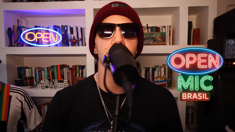 Rapper de gorro vermelho e óculos escuros canta ao microfone diante do letreiro neon do Open Mic Brasil