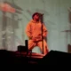 Anderson .Paak canta de moletom laranja, com microfone no pedestal, em show de dezembro de 2018