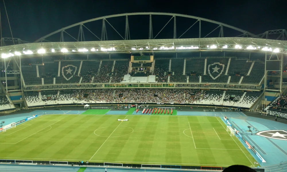 Vista interna do Estádio Nilton Santos, o Engenhão, à noite, com gramado e arquibancadas iluminados