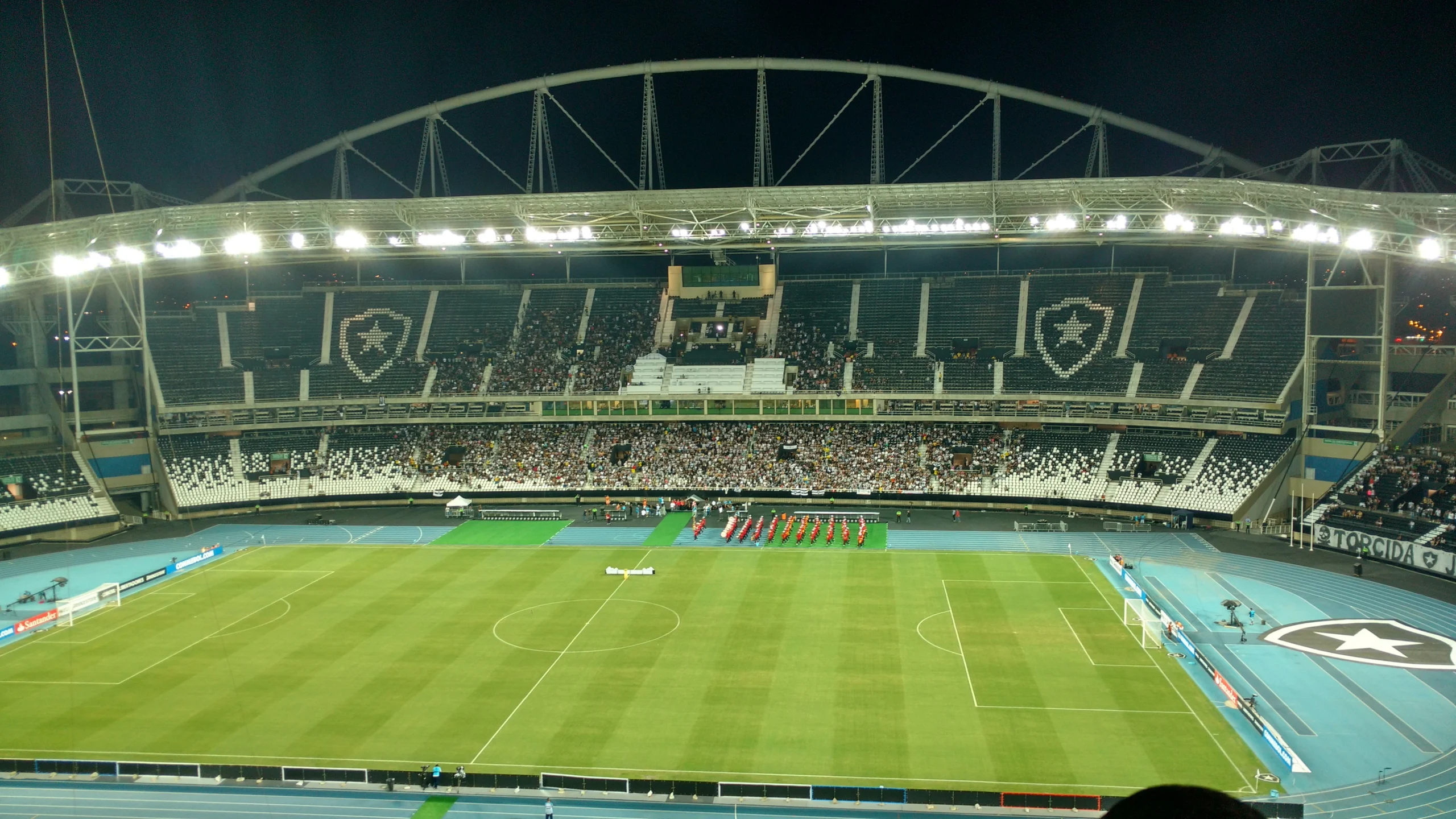 Vista interna do Estádio Nilton Santos, o Engenhão, à noite, com gramado e arquibancadas iluminados