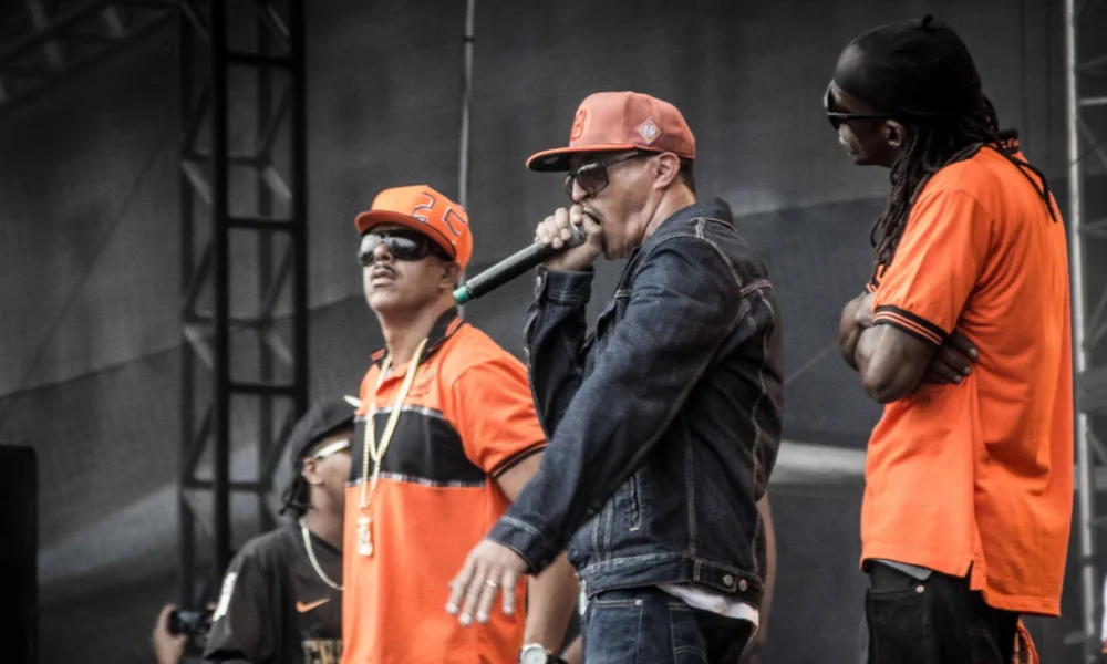 Integrantes dos Racionais MC's no palco da Virada Cultural de São Paulo, com camisas laranja e jaqueta jeans
