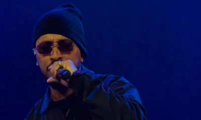 Rashid de gorro e óculos escuros canta ao microfone sob luz azul no show Cumulonimbus