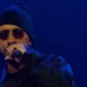 Rashid de gorro e óculos escuros canta ao microfone sob luz azul no show Cumulonimbus