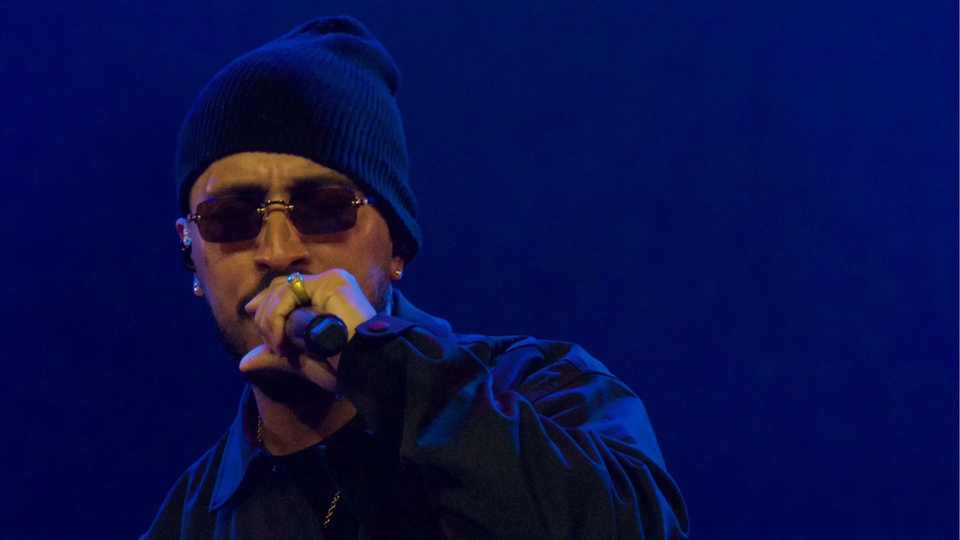 Rashid de gorro e óculos escuros canta ao microfone sob luz azul no show Cumulonimbus