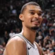 Victor Wembanyama sorri em quadra com o uniforme branco do San Antonio Spurs